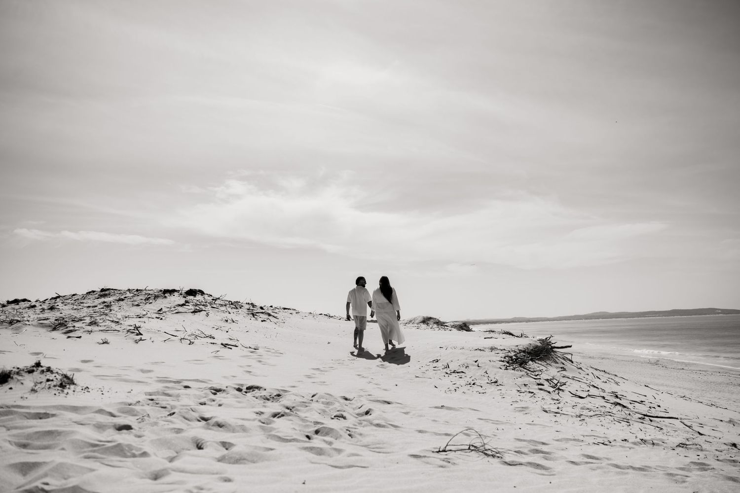 Two figures walking together on a sandy beach dune overlooking the ocean in black and white.