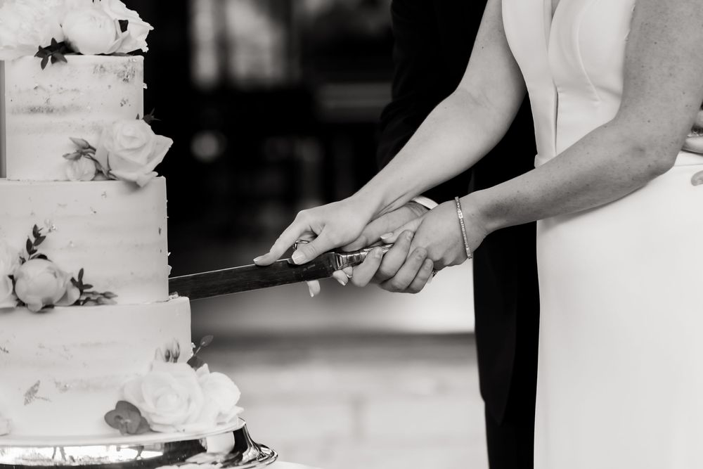 Black and white close-up of hands cutting a tiered wedding cake.