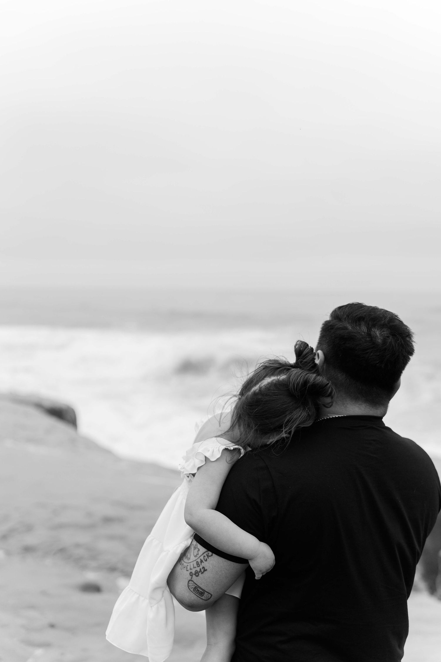 A tender black and white moment of a couple embracing while looking out at the ocean waves.