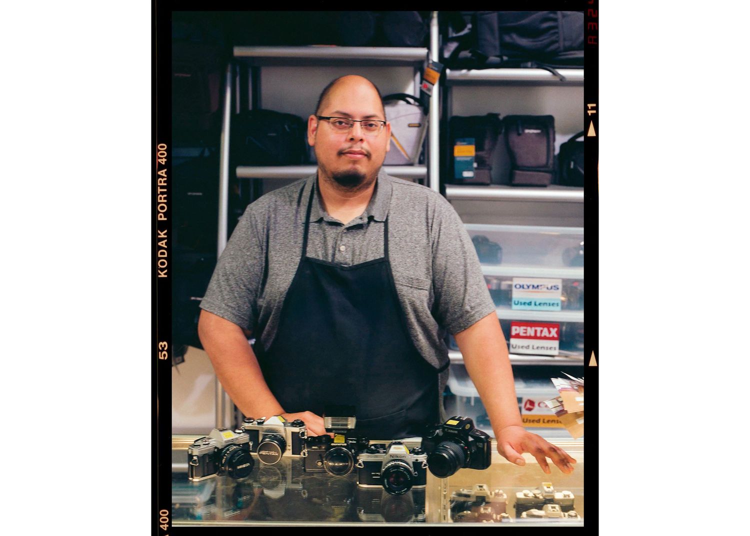 Camera repair technician standing behind a workbench with vintage film cameras in a photography shop.
