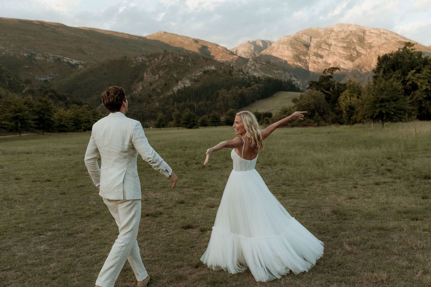 Bride in white tulle gown and groom in ivory suit dance in a mountain meadow at golden hour.