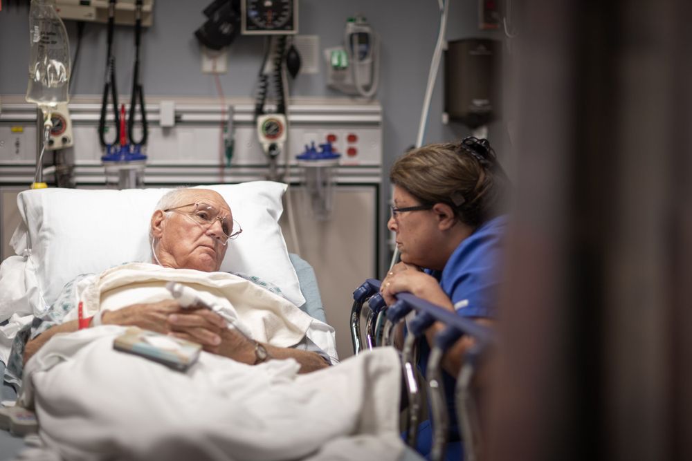 Medical professional in blue scrubs consults with patient lying in hospital bed with medical equipment in background.