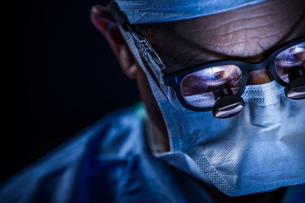 Close-up view of a surgeon wearing surgical mask and protective glasses during operation in blue-tinted lighting.
