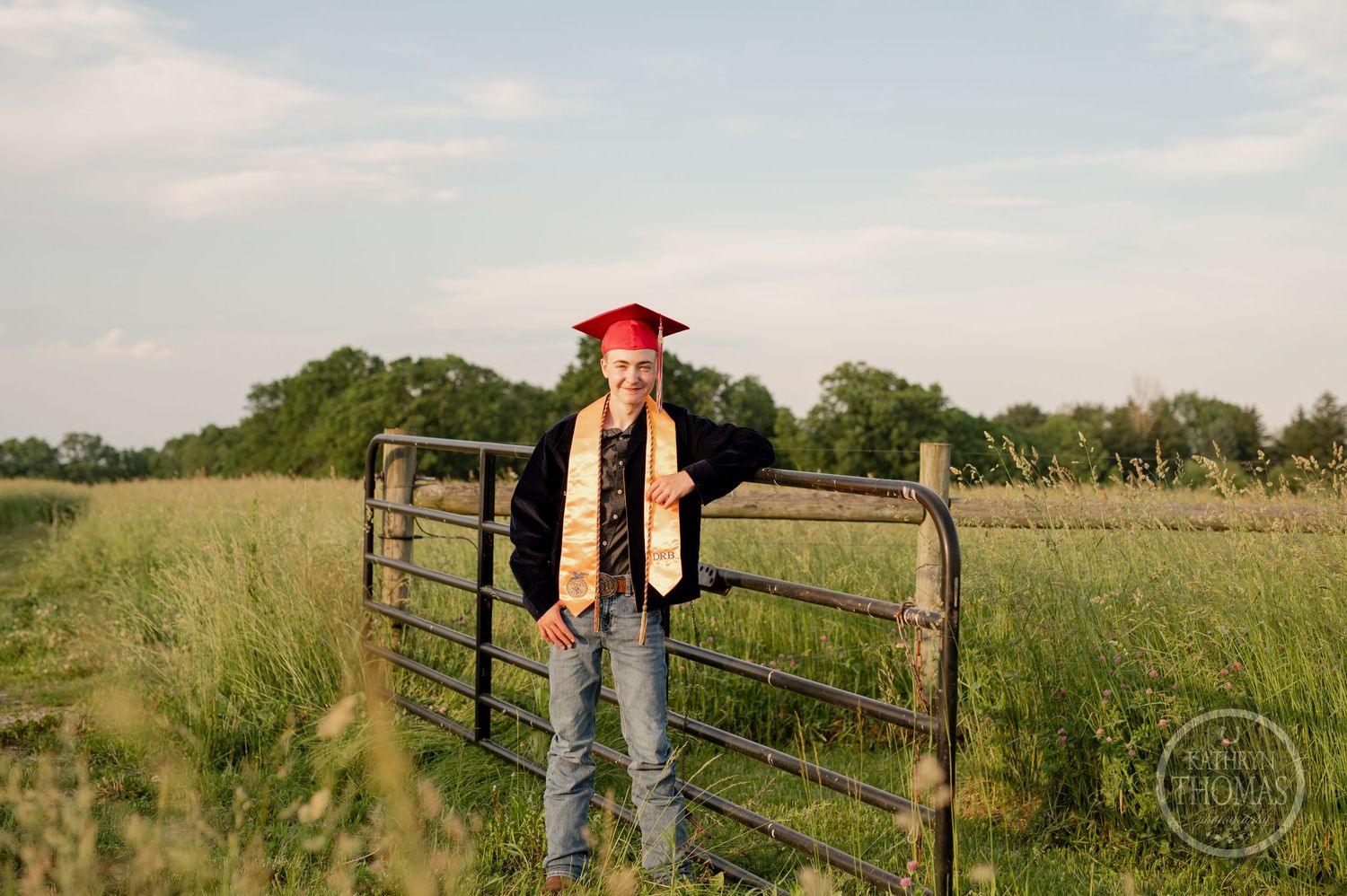 Graduate in red cap and FFA stole stands by rustic farm gate in grassy field.