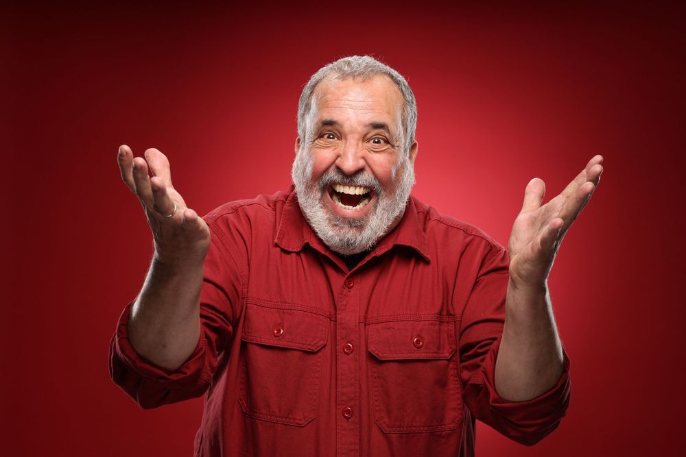 A joyful person wearing a red button-down shirt raises their hands with a big laugh against a red background.