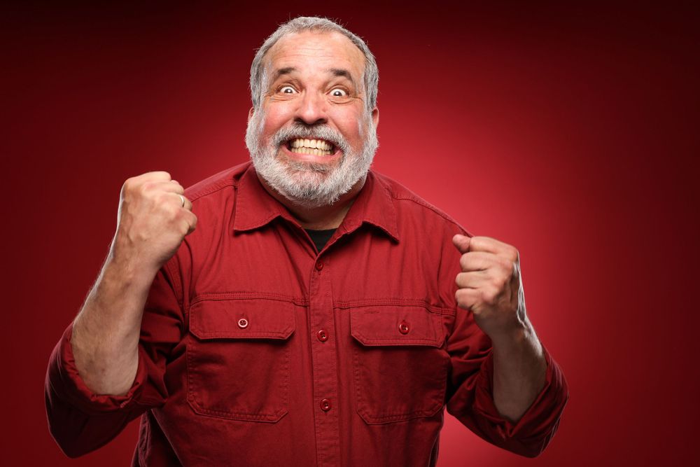 Excited older man wearing red shirt making celebratory gesture with fists against red background.