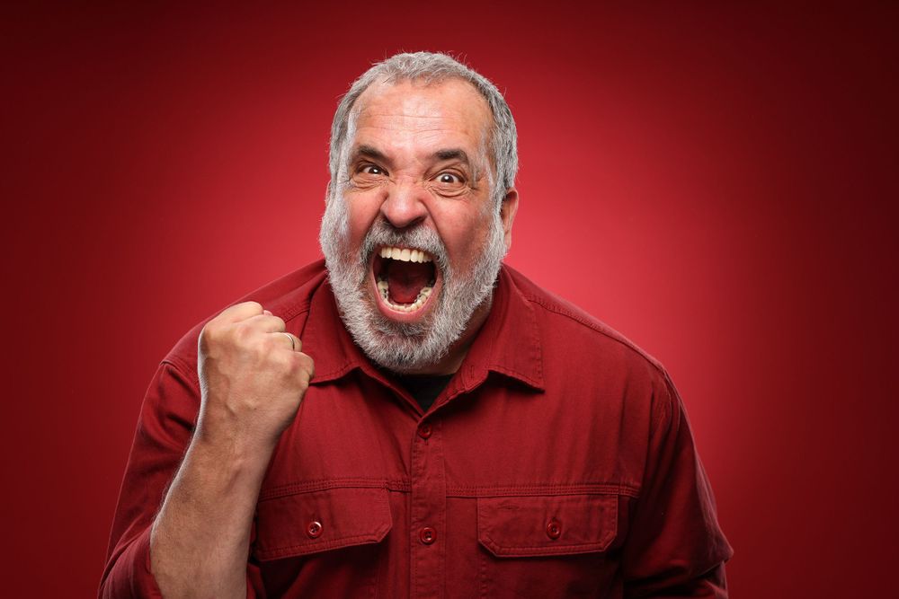 A person with a grey beard wearing a red shirt gestures angrily against a dark red background.