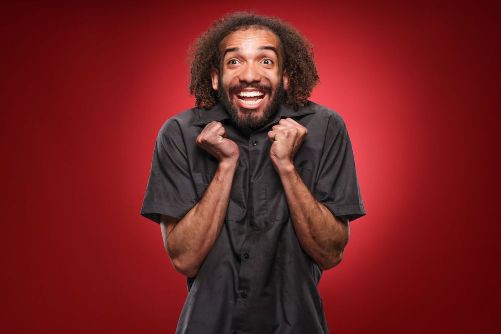 Excited person with curly hair and black shirt making joyful expression against red background.