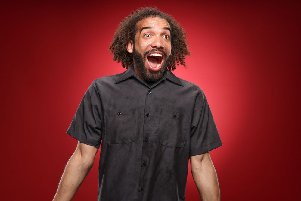 Excited person with curly hair and black button-up shirt expresses joy against red background.