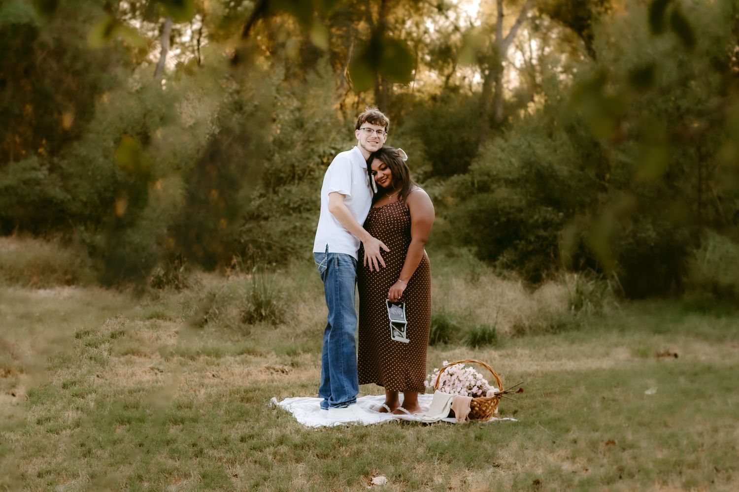 A couple stands together in a meadow at sunset with a picnic basket and blanket spread on the ground.