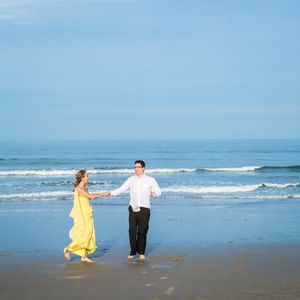 Un couple, l'une en robe jaune et l'autre en chemise blanche, danse ensemble sur une plage avec des vagues déferlantes et un ciel bleu.