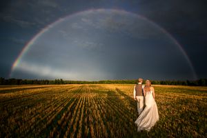 Photo extraordinaire d'un couple de mariés marchant dans un champ de blé doré sous un magnifique arc-en-ciel durant un coucher de soleil.