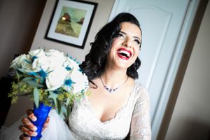Une mariée heureuse en robe de mariée en dentelle blanche pose avec un bouquet de fleurs bleues et blanches dans un décor intérieur.