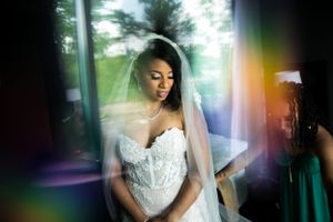 Une jolie femme noire, vêtue d'une robe de mariée en dentelle, pose près d'une fenêtre où l'éclairage et le voile créent un effet éthéré.