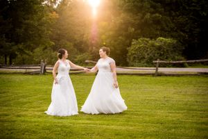 Deux femmes lesbiennes en robes de mariée blanches se tiennent la main en marchant dans un champ baigné de soleil à l'heure dorée.