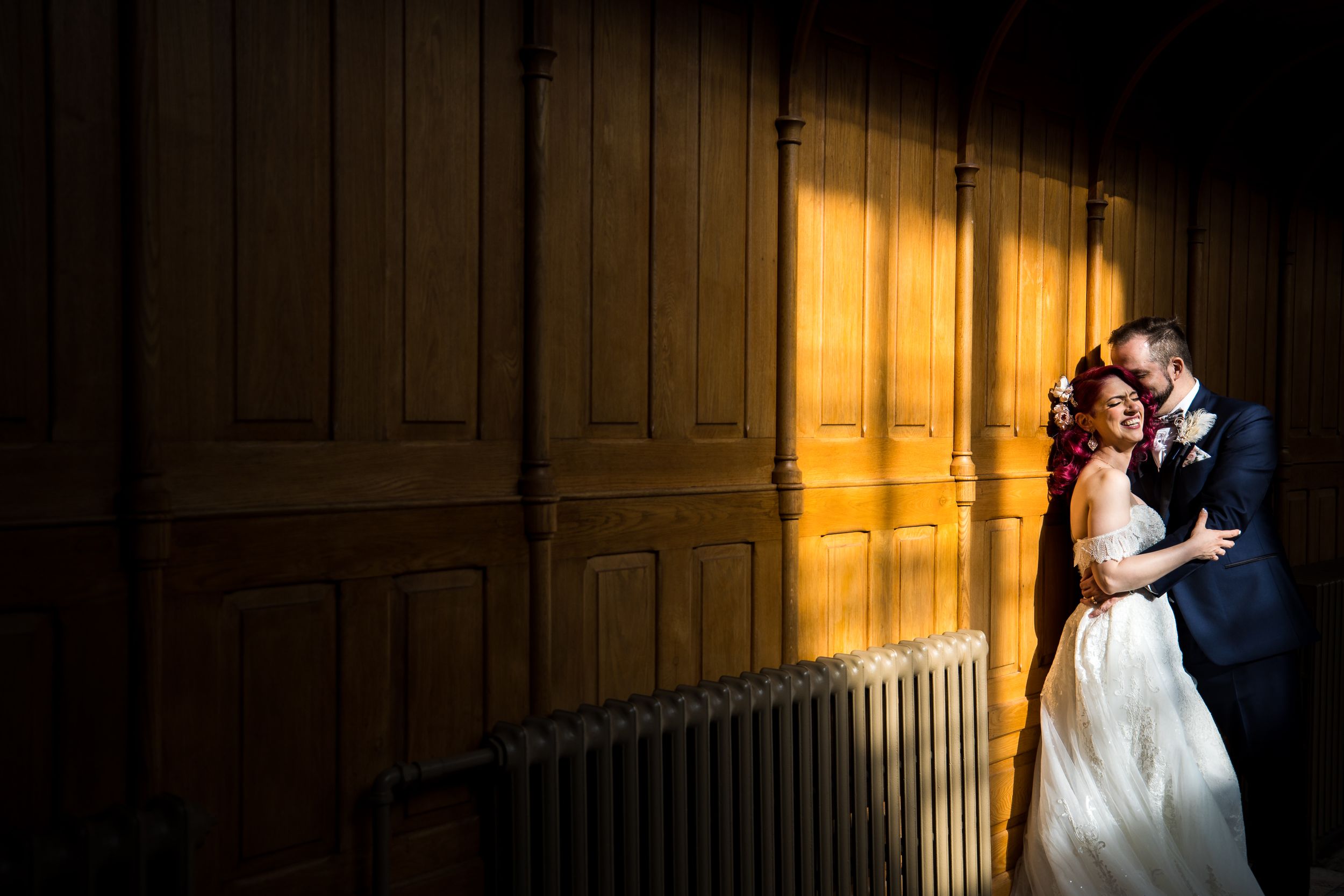 À l’Abbaye d’Oka, un couple partage un moment intime de mariage contre un mur en bois chaleureusement éclairé, avec des ombres dramatiques.