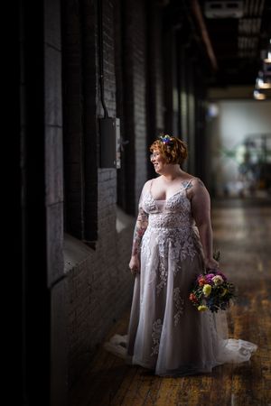 À L’Ambroisie et L’Espace Canal à Montréal, une mariée en robe de dentelle, tenant un bouquet coloré, se tient dans un couloir rustique.