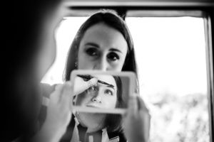 Photo en noir et blanc montrant une mariée qui se regarde dans un miroir pendant qu’elle se fait maquiller, à l’hôtel Rive-Gauche de Belœil.