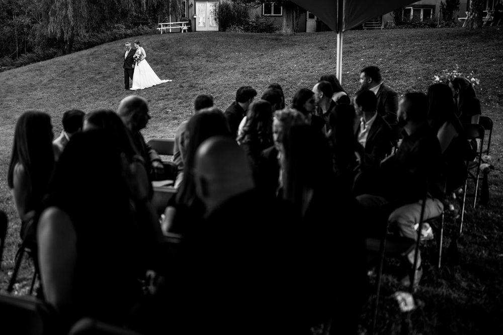 Photo en noir et blanc d’une cérémonie de mariage où les invités en premier plan regardent la mariée marcher avec son père en arrière-plan.