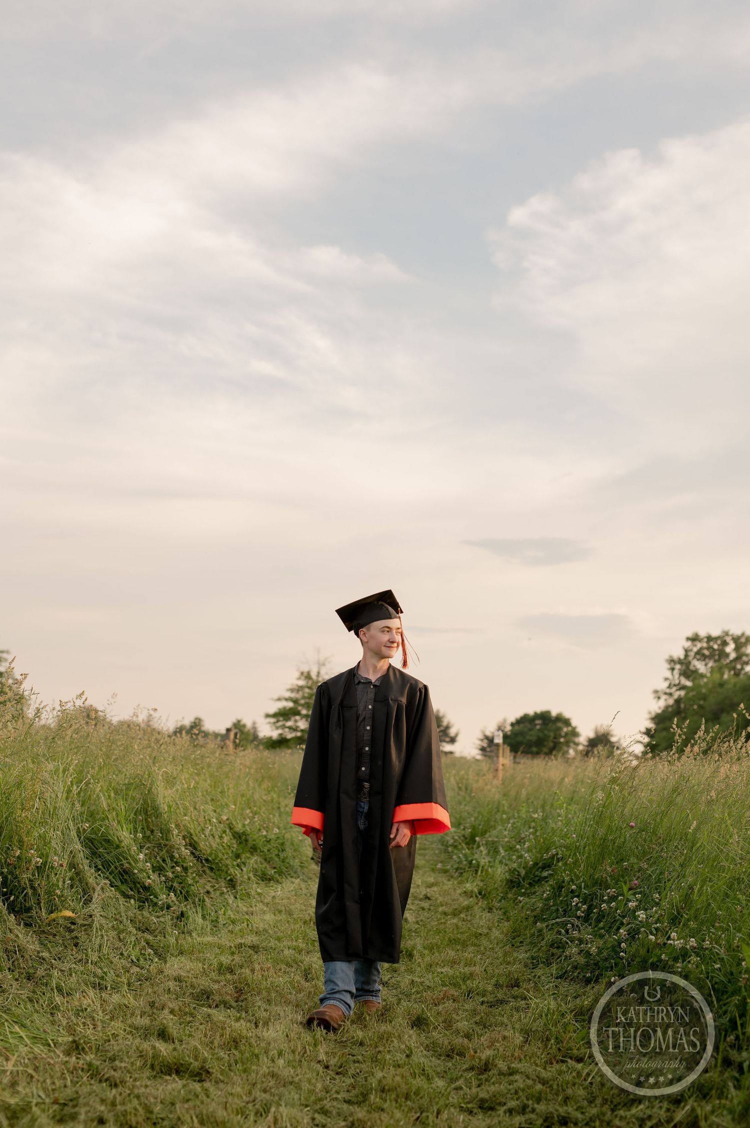 Graduate walks along path through meadow wearing traditional black graduation gown.