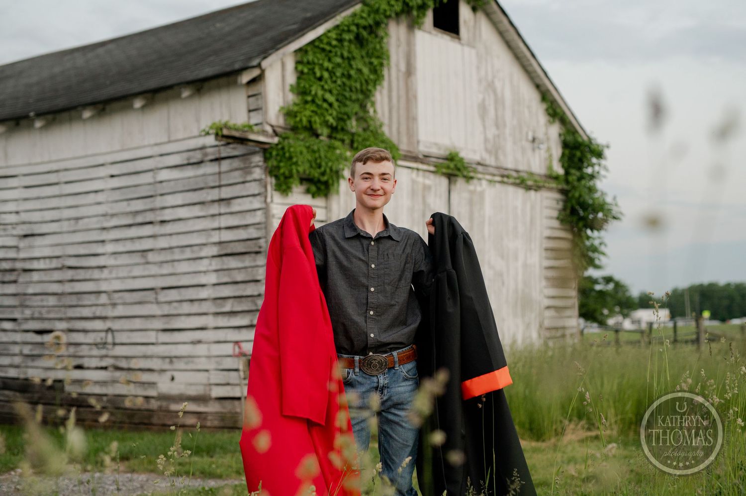 Graduate stands with red and black gowns in front of rustic white barn with ivy.