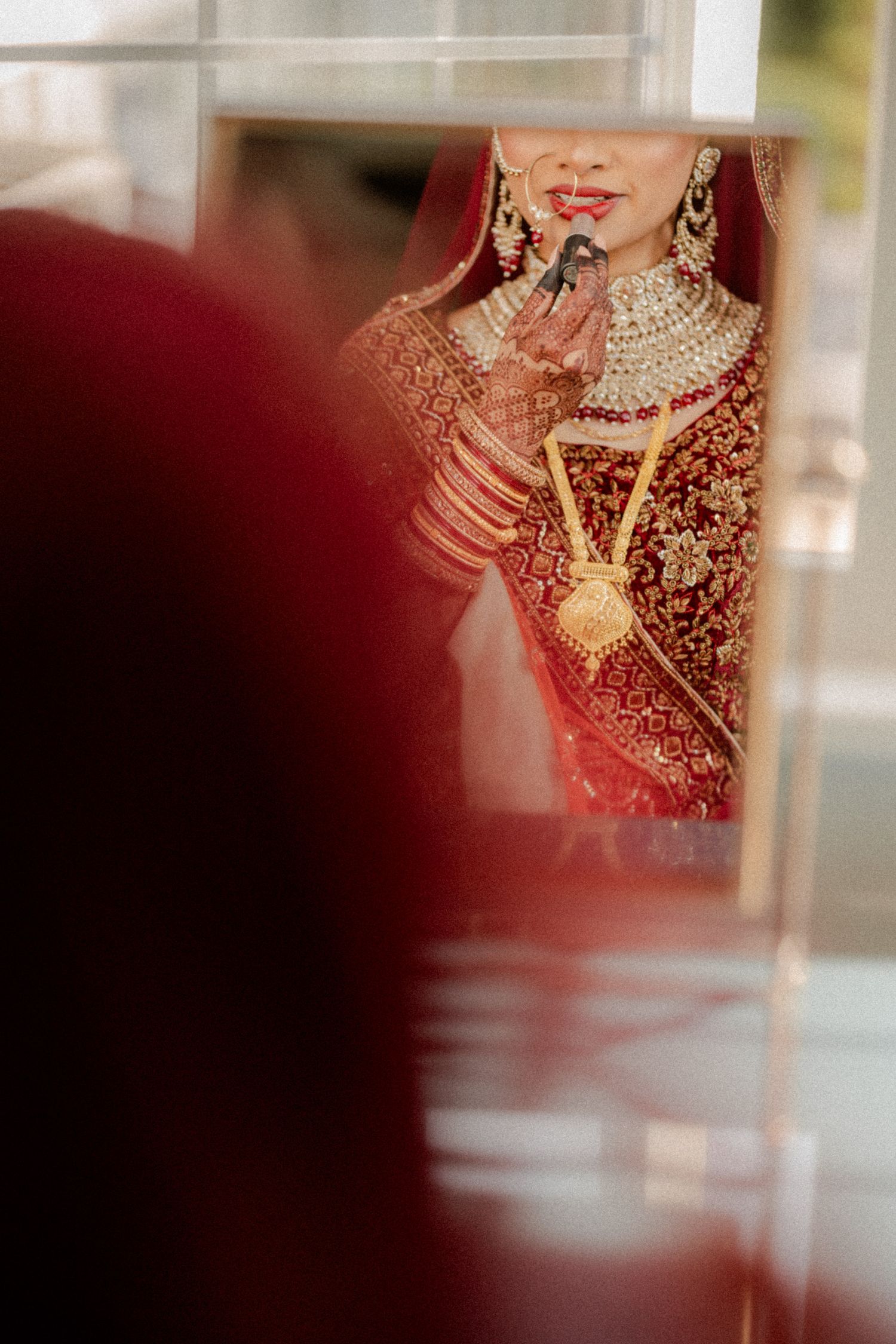 Through a soft blur, a glimpse of intricate red and gold bridal attire with ornate jewelry and traditional Indian wedding details.