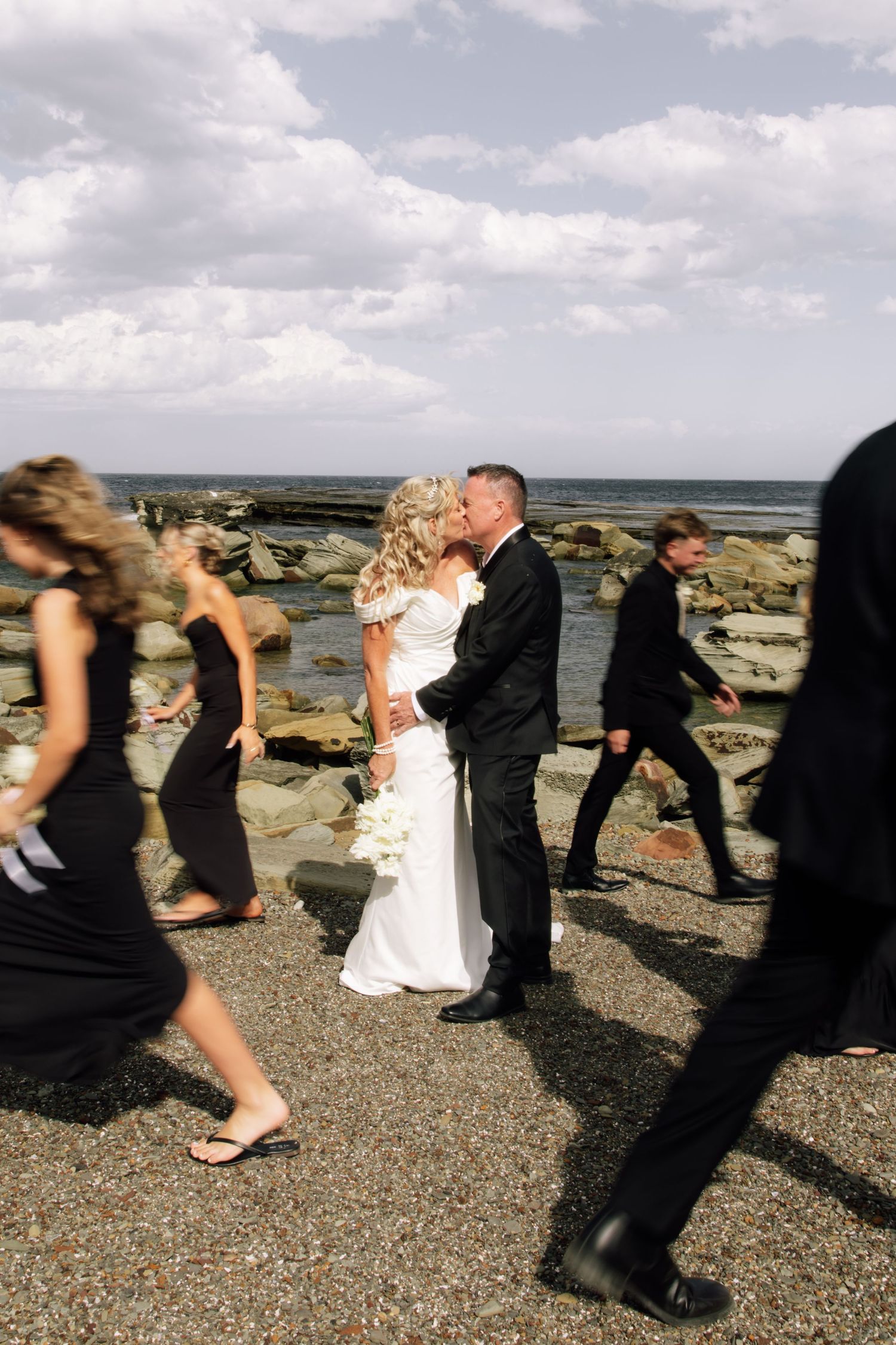 Wedding party walks in line along rocky beach shore with dramatic cloudy sky in background.