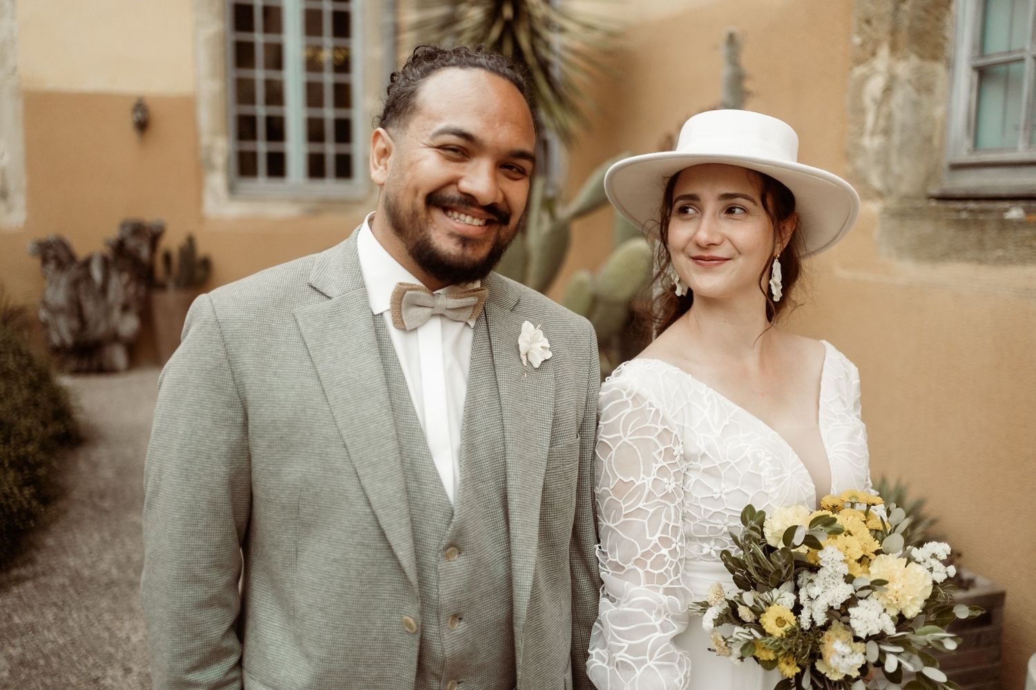 Newlywed couple poses for wedding portraits in front of a beige stone building with white flowers.