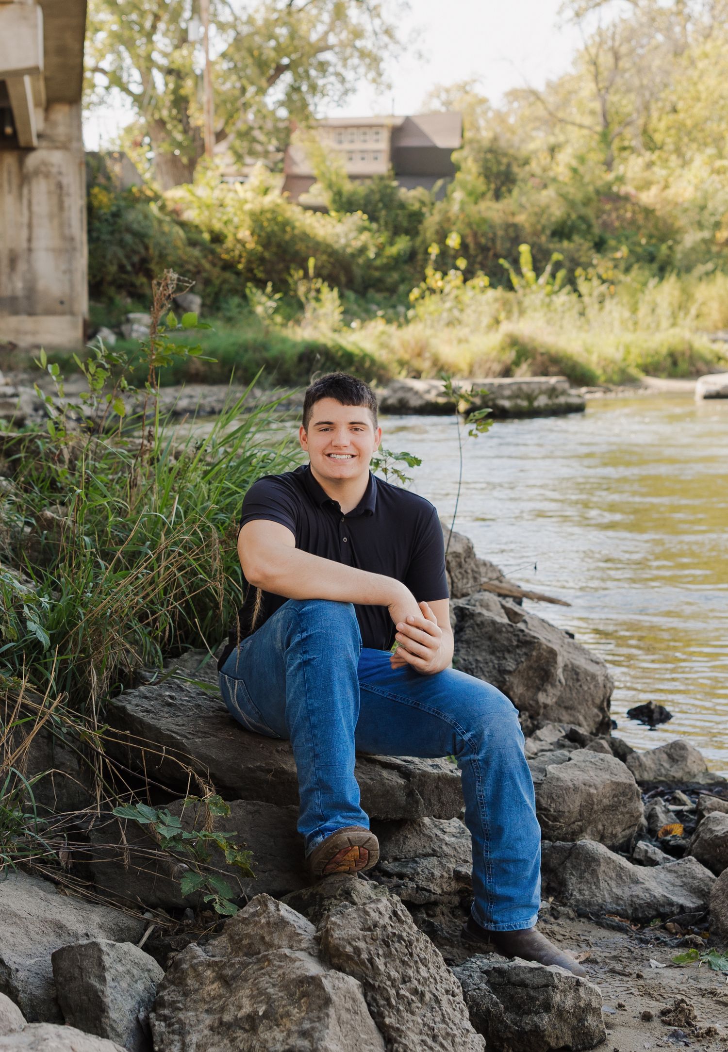 A person sits casually on rocky riverbank wearing jeans and black shirt with flowing water and greenery in background.