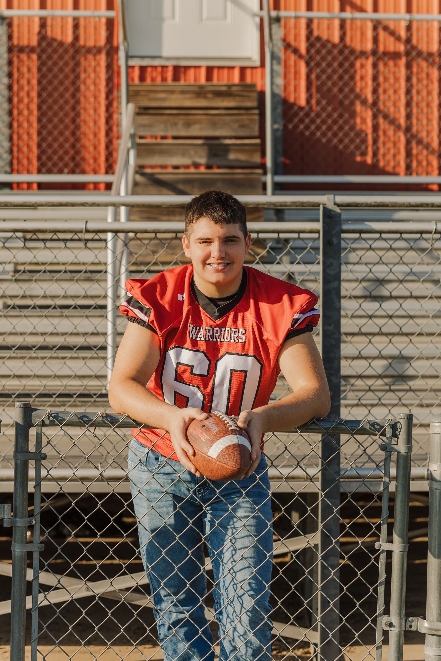 Football player in red jersey and jeans poses on bleachers during outdoor photo session.