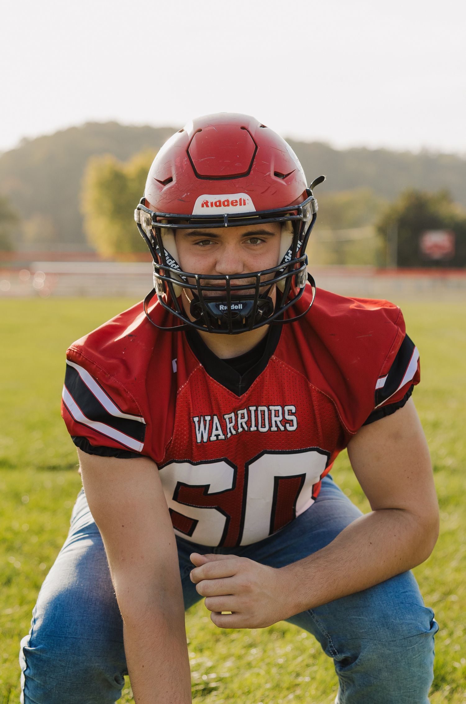 High school football player in red Warriors uniform and jeans poses on field during golden hour.