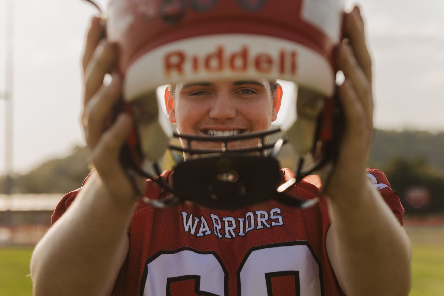 Football player in Warriors uniform holds Riddell helmet against sunset backdrop.
