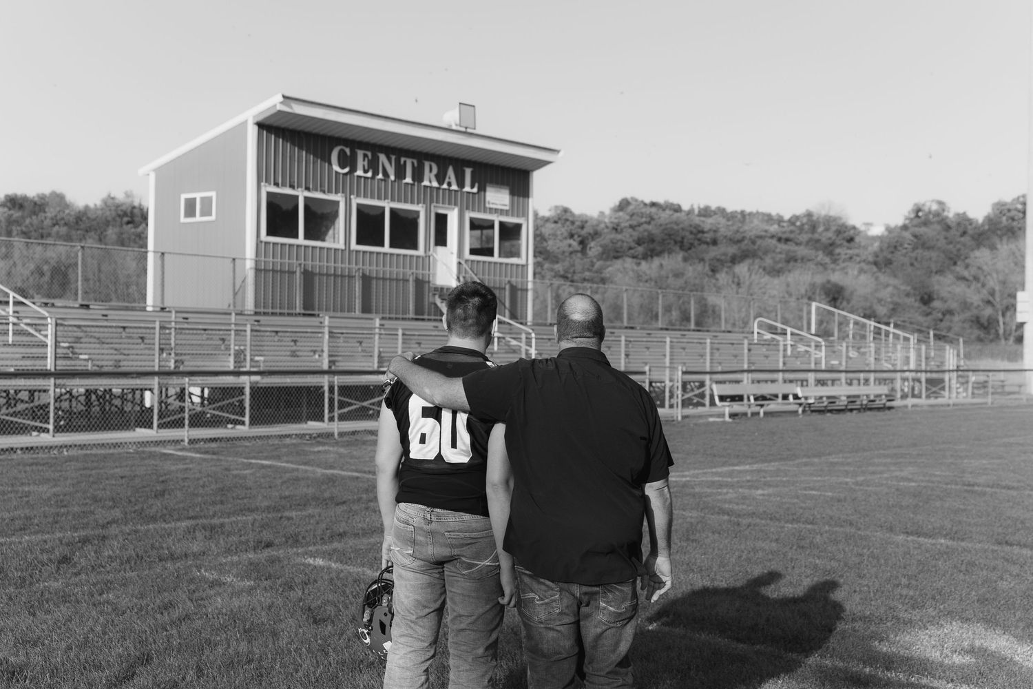 Two figures stand together looking at a football field with a press box and bleachers in black and white.