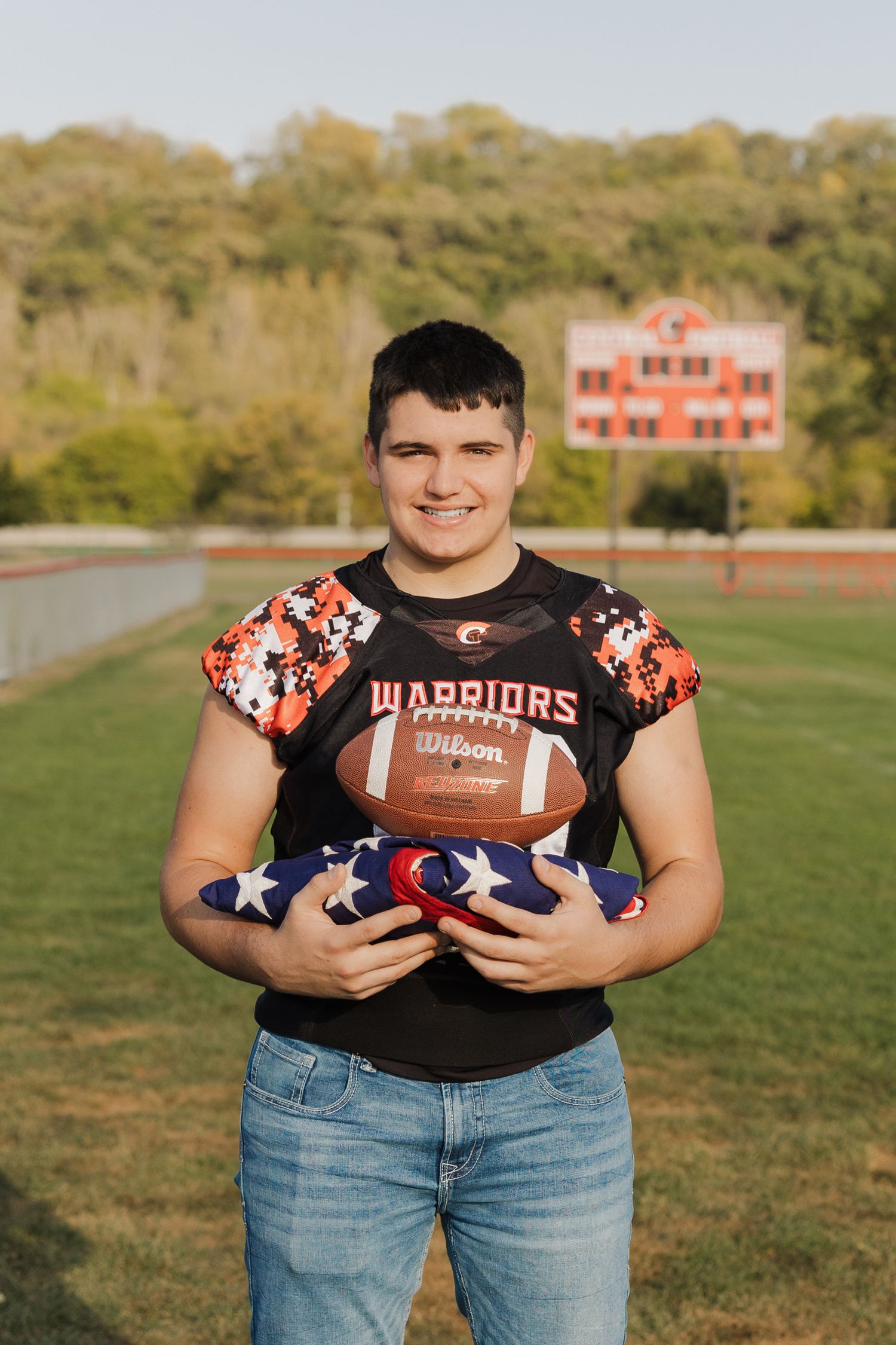 Football player in Warriors jersey holds a football and American flag on a sunny field with scoreboard in background.