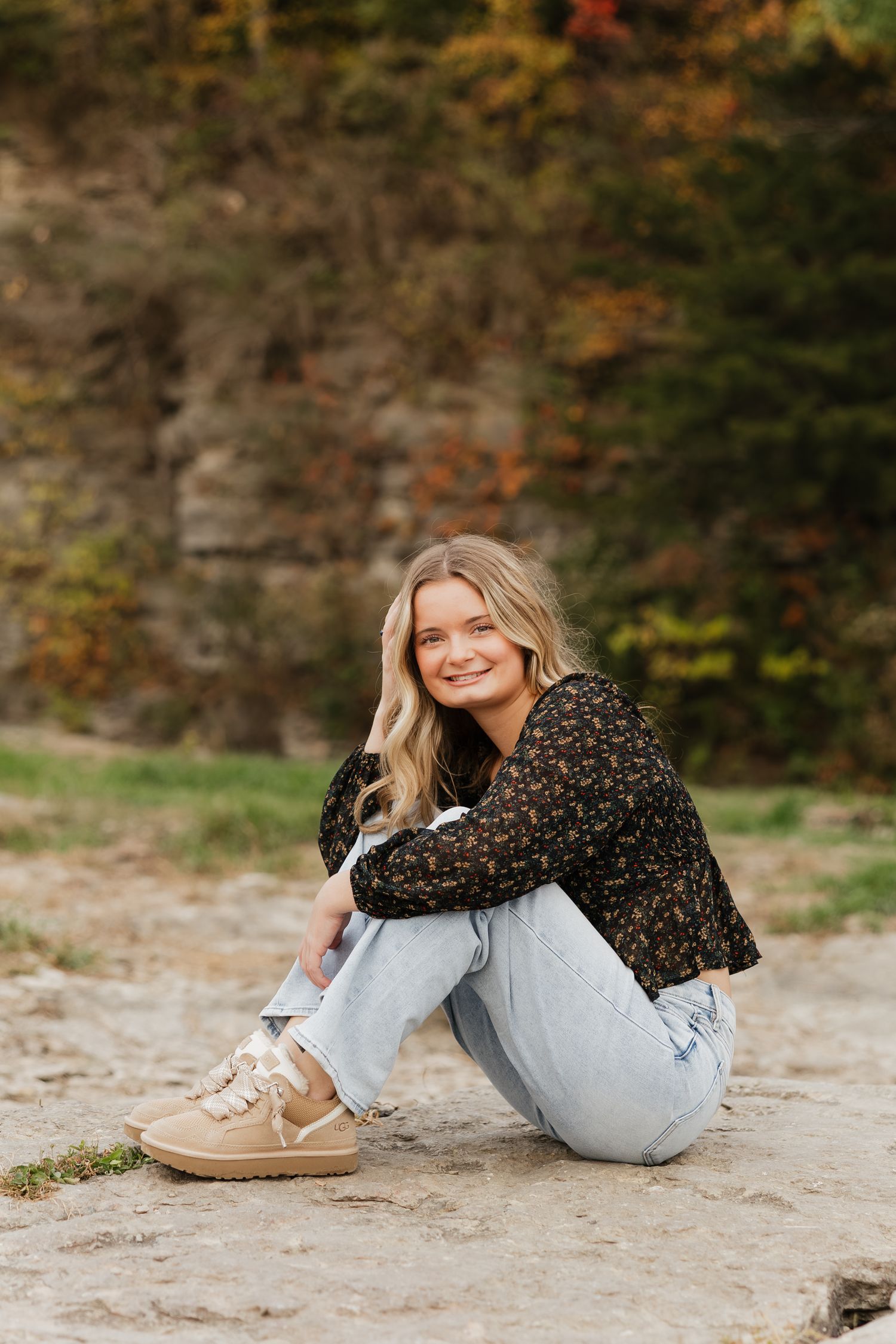 A person in a floral top and light jeans relaxes on a sandy ground during an autumn outdoor photoshoot.