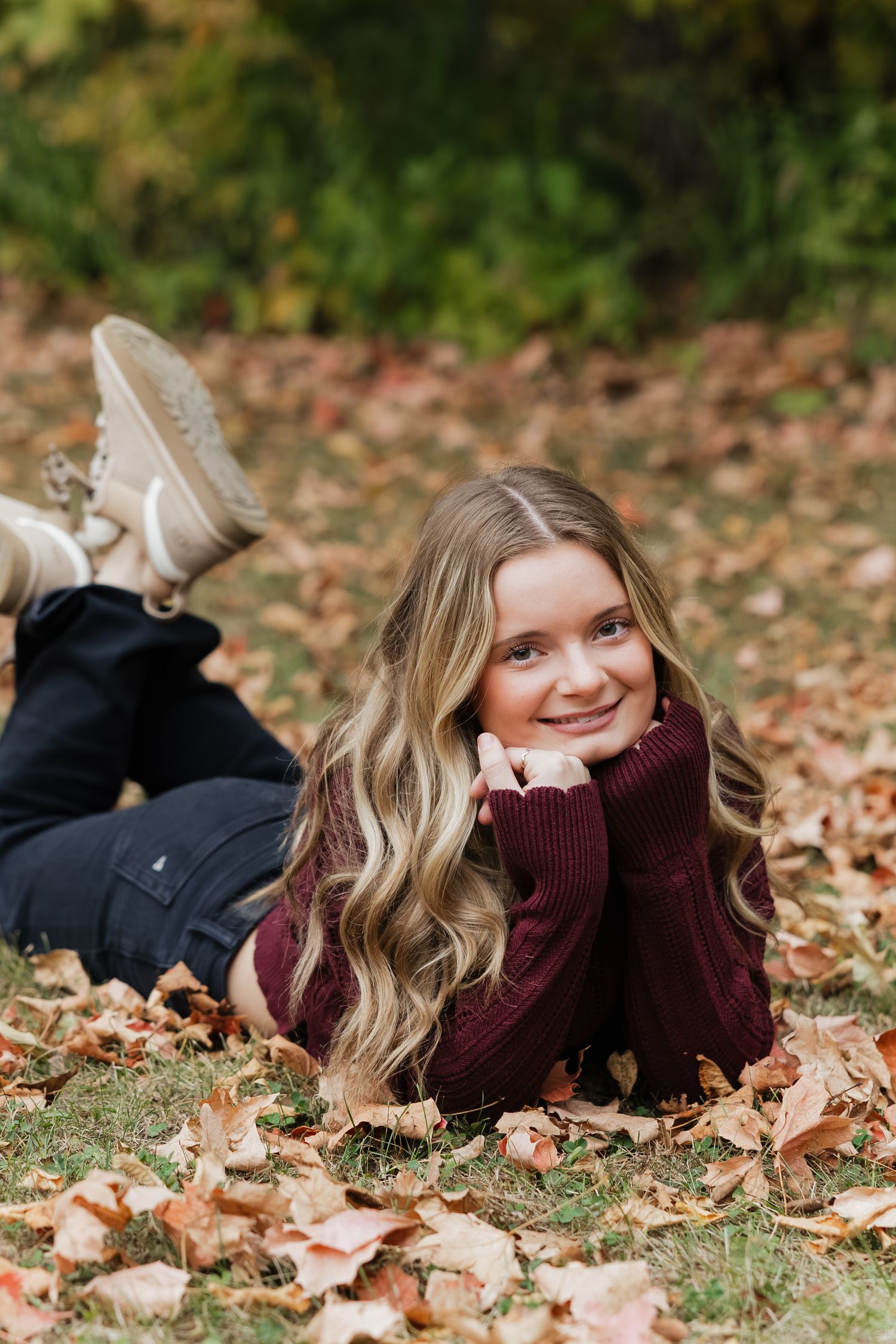 A person in a burgundy sweater lies on autumn leaves while smiling at the camera during a fall photo session.