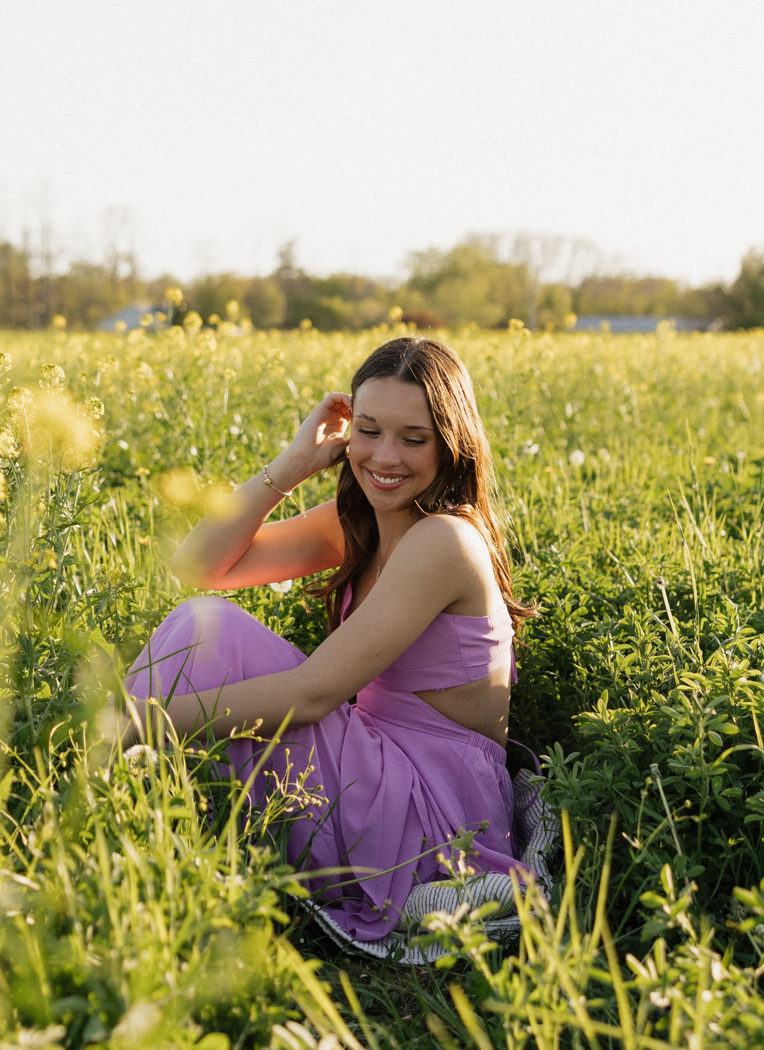 A person in a purple dress sits among yellow wildflowers in a sunny field during sunset.