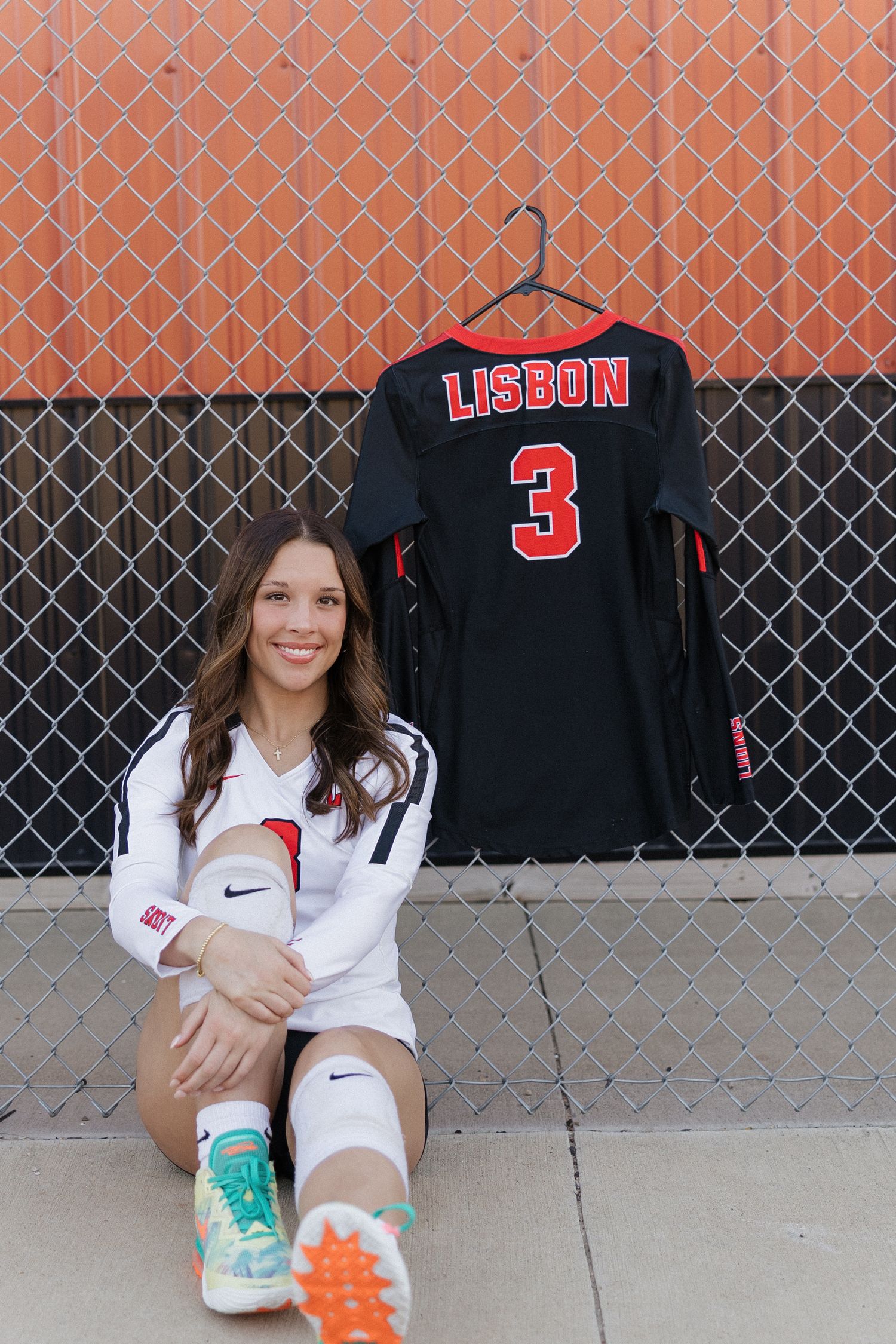 Student athlete poses against chain link fence wearing white polo and colorful sneakers with Lisbon jersey displayed.