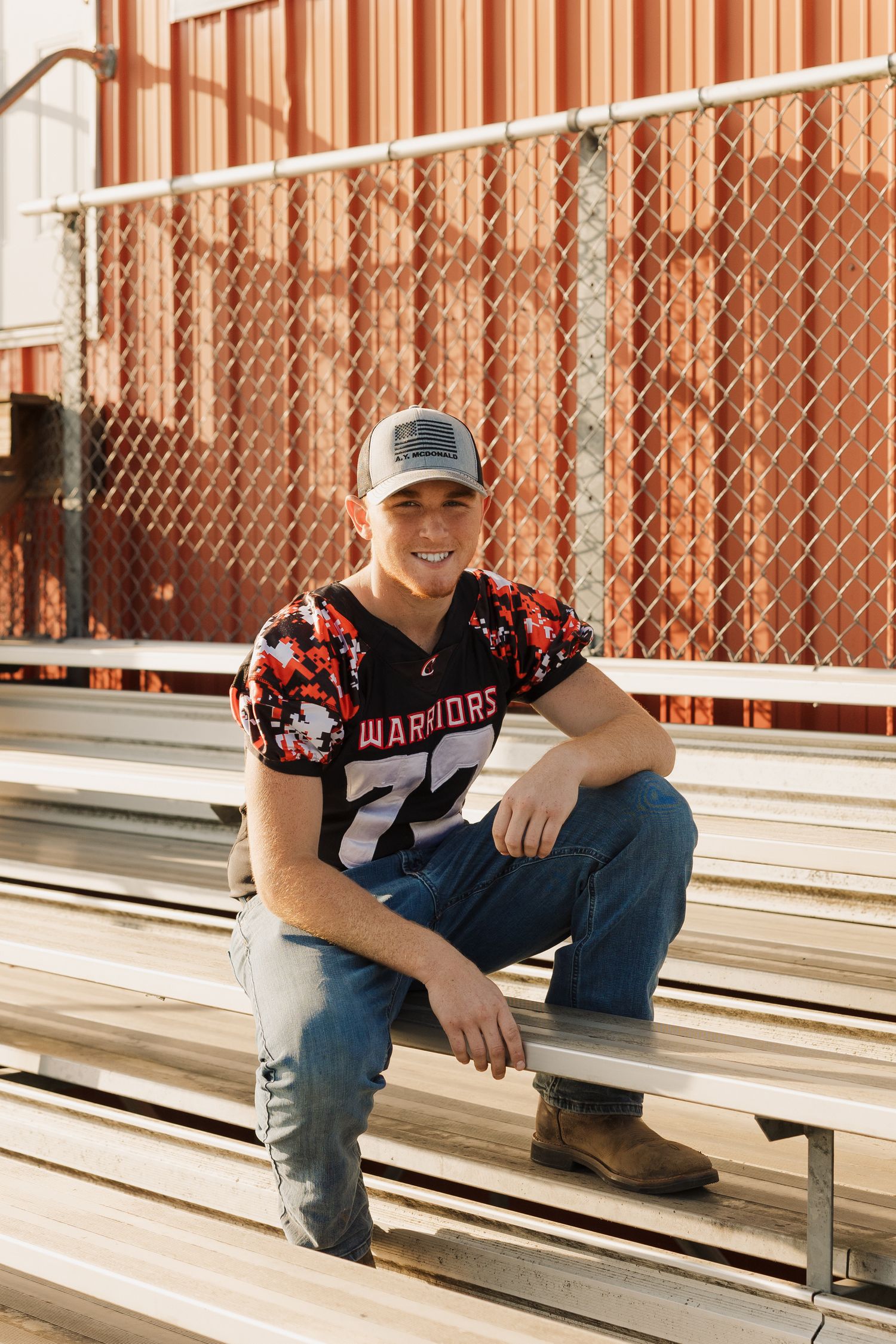 Young athlete in a Warriors jersey and baseball cap sits casually on metal bleachers in warm sunlight.