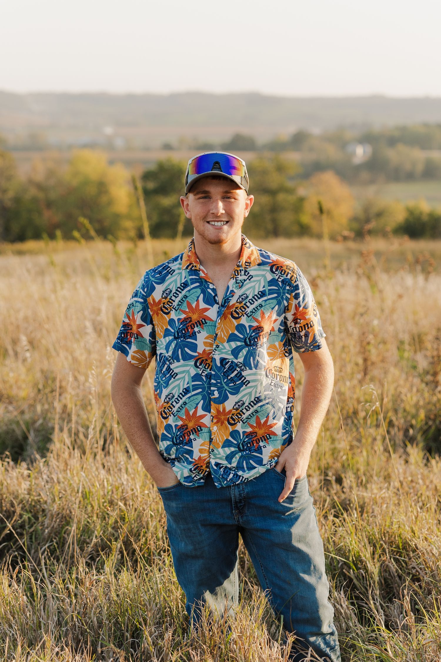 A person in a tropical print shirt and jeans sits in a golden field at sunset.