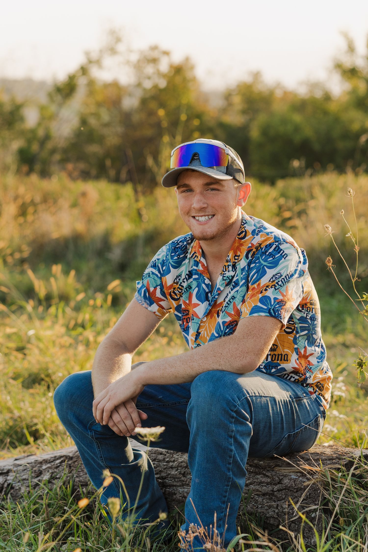 Person in colorful Hawaiian shirt and baseball cap sits casually in a grassy field during golden hour.