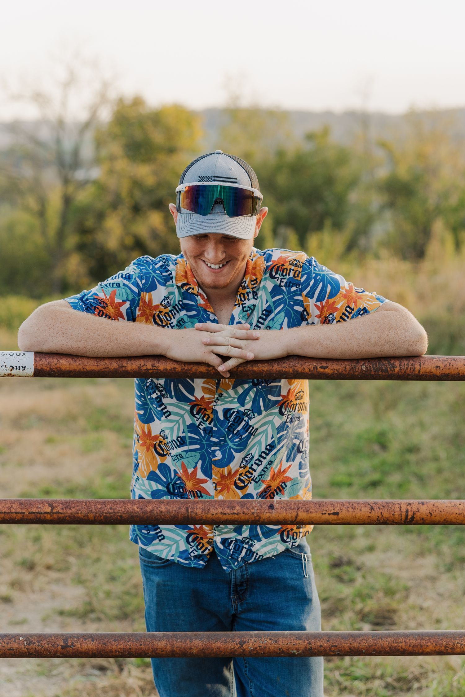 Person in vibrant floral shirt leans on rustic wooden fence while smiling outdoors.