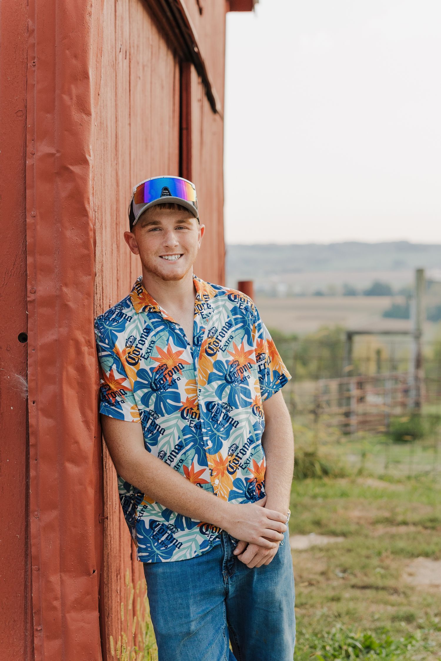 Someone wearing a colorful tropical print shirt and baseball cap poses against a rustic red barn door in a rural setting.