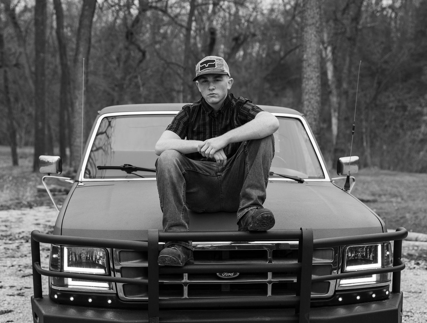 A black and white photo series showing someone posing with a classic muscle car in a wooded setting.