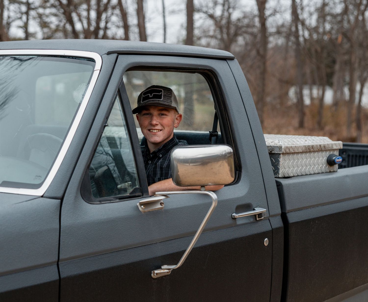 A person smiles from the driver's seat of a vintage gray pickup truck with chrome side mirrors.