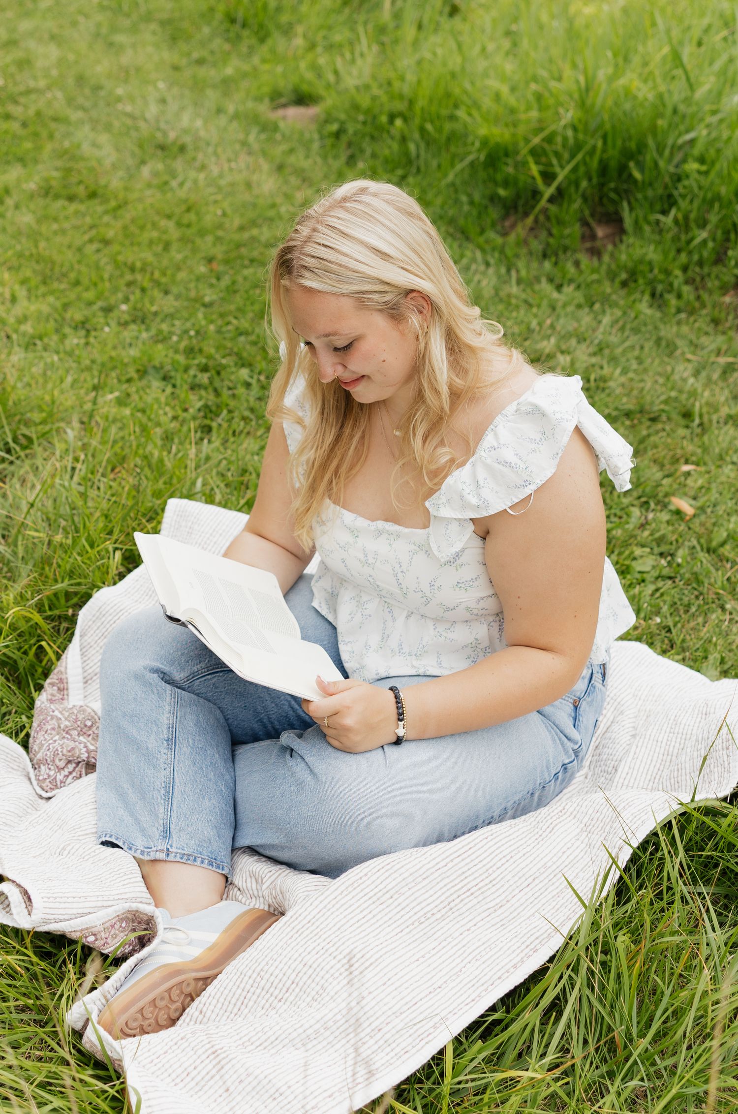 Someone reads a book while relaxing on a white blanket in the grass wearing a white top and jeans.