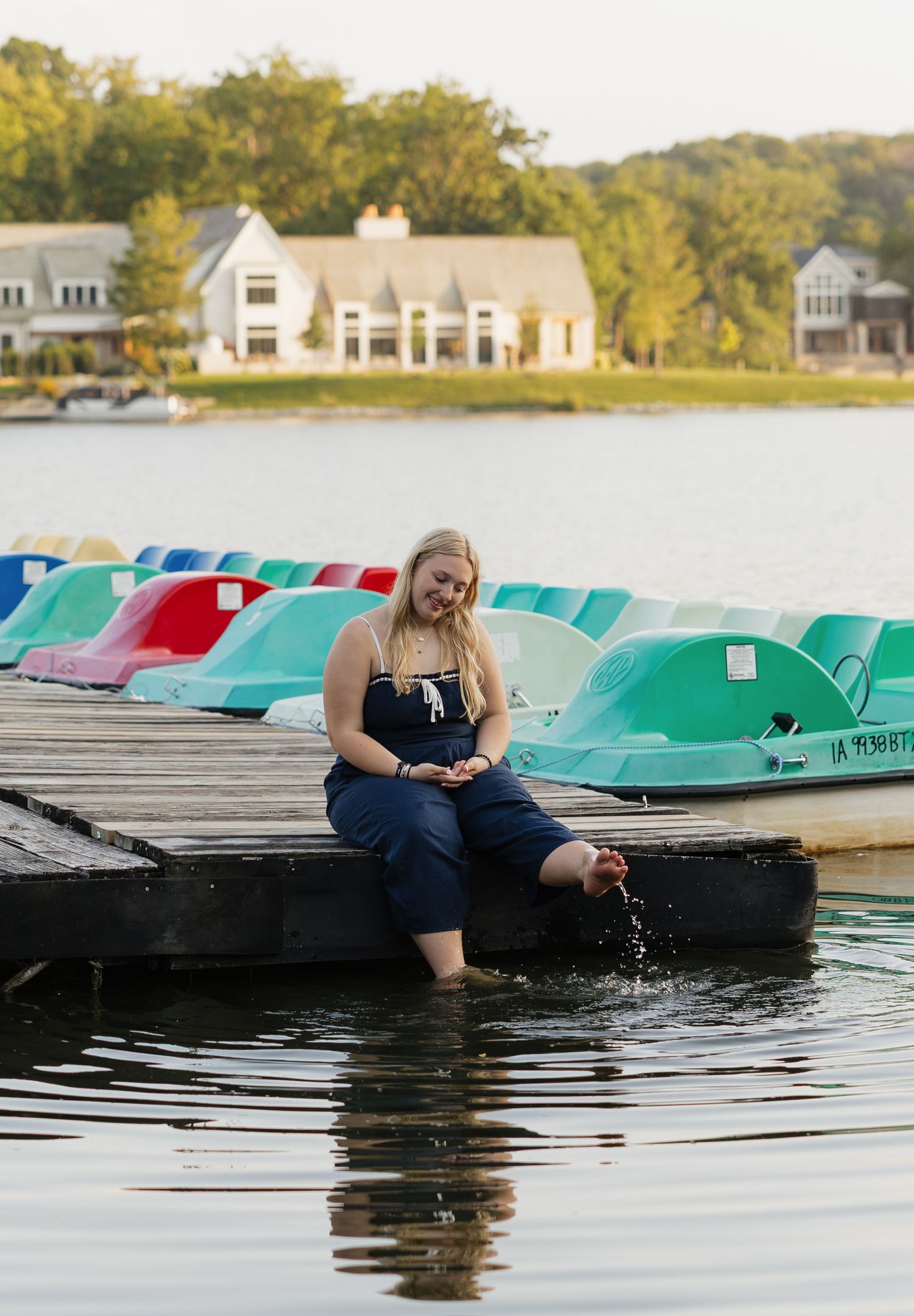 Someone sits on a dock by a lake with colorful pedal boats lined up, waterfront homes visible in the background.