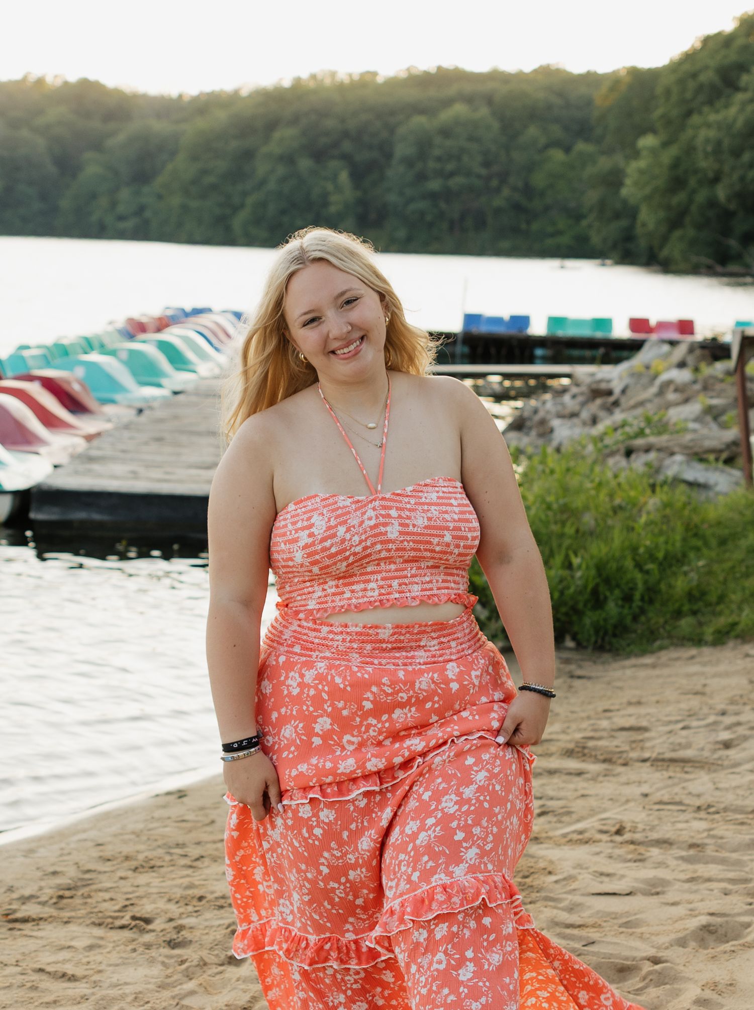 A person in a coral floral sundress poses outdoors near a lake with colorful boat rentals in the background.