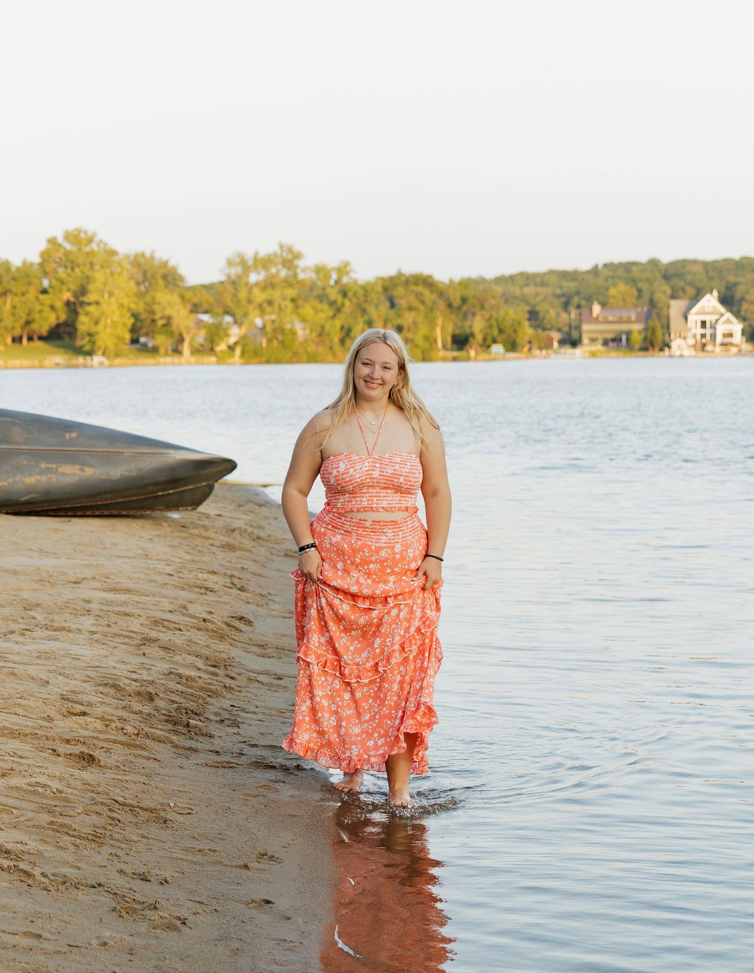 A person wearing a coral floral dress stands in shallow water at the lake's edge during golden hour.