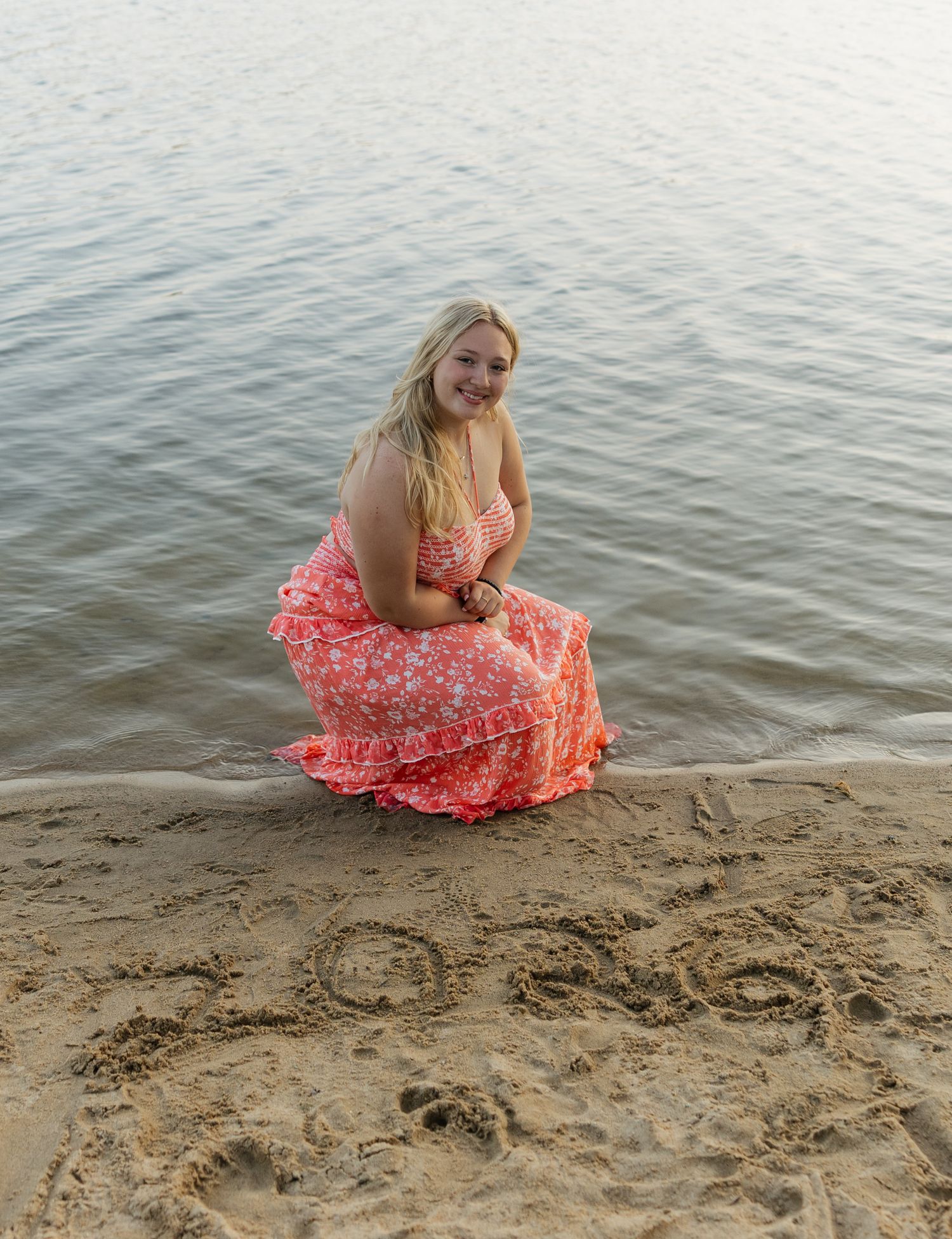 A person in a coral floral dress sits by the water's edge on a sandy beach at sunset.